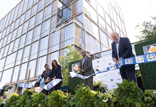 Four adults on a stage cut a ribbon in front of a large glass building